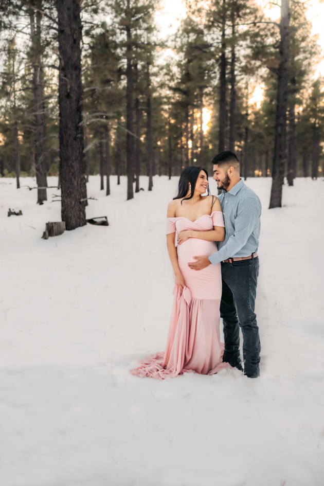 Pregnant woman in an off the shoulder pink gown with husband holding her in the snow in Flagstaff, Arizona