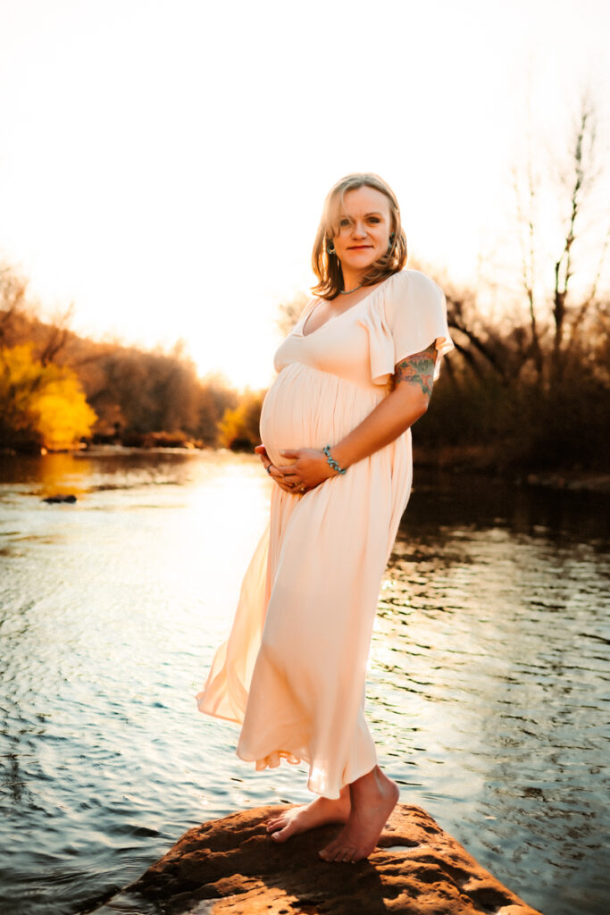 Expecting mom holding her belly in a maxi dress at the creek in Sedona.